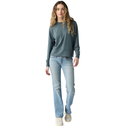 A person with long black hair is standing against a plain white background, wearing a Cozy Earth Women's StudioLite Crewneck in light green and dark green pants. They are looking down with a gentle smile, gently touching their neck with one hand. |Color:Haze