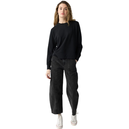 A person with long black hair is standing against a plain white background, wearing a Cozy Earth Women's StudioLite Crewneck in light green and dark green pants. They are looking down with a gentle smile, gently touching their neck with one hand. |Color:Haze