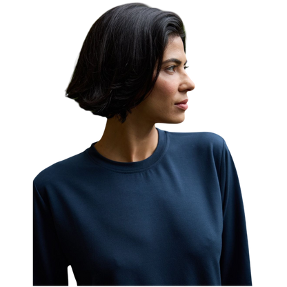 A person with long black hair is standing against a plain white background, wearing a Cozy Earth Women's StudioLite Crewneck in light green and dark green pants. They are looking down with a gentle smile, gently touching their neck with one hand. |Color:Haze