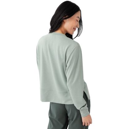 A person with long black hair is standing against a plain white background, wearing a Cozy Earth Women's StudioLite Crewneck in light green and dark green pants. They are looking down with a gentle smile, gently touching their neck with one hand. |Color:Haze