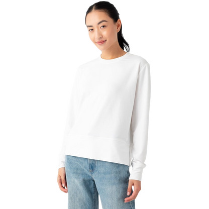 A person with long dark hair, wearing the Women's StudioLite Crewneck from Cozy Earth and light blue jeans, is standing against a plain white background. They are slightly smiling and have one hand in their pocket. |Color:White